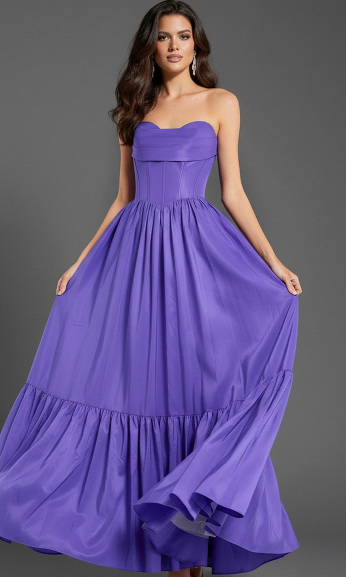A woman stands holding the sides of the Strapless Long Ruffled Prom Dress: Jovani 47883, featuring a fitted corset-style bodice and flared purple skirt, set against a plain gray background.